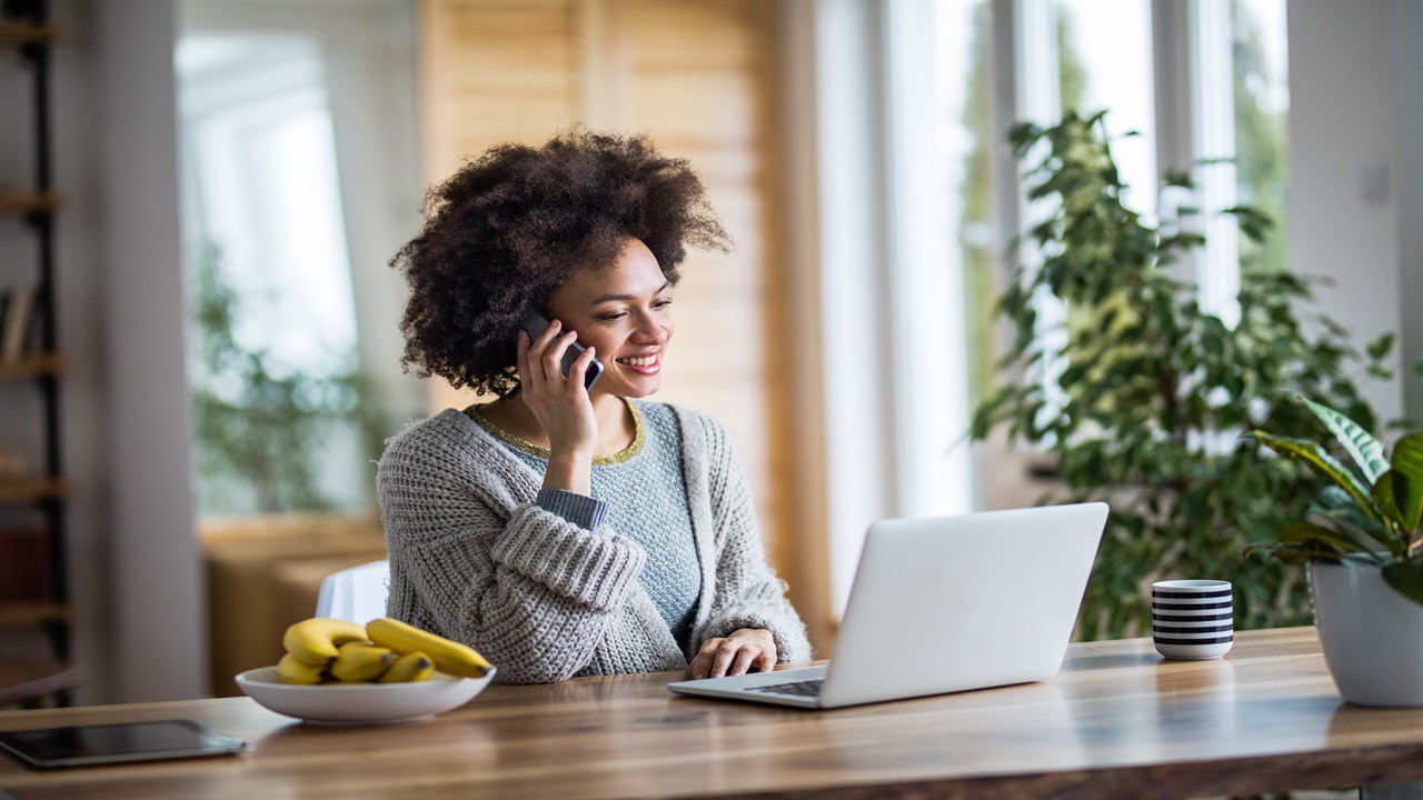 Happy African American woman talking to someone over mobile phone at home while using laptop.